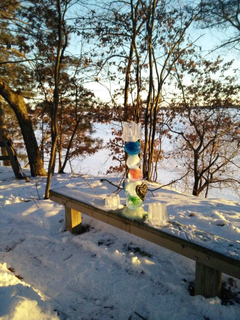 A scene from the edge of the labyrinth looking out at the frozen snow-covered lake through the trees at a previous gathering. An Ice sculpture of a tower of 7 pieces, 5 of them round balls and several colored, sits on a wooden bench.